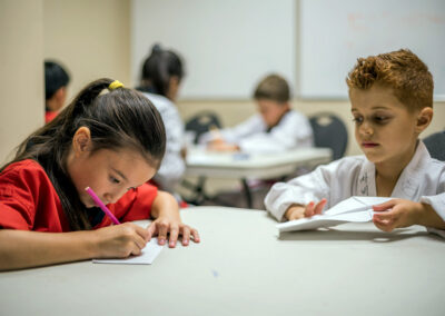 Two students work at a classroom table: girl in red foreground writes with a pink pencil while a boy in a white uniform looks at his paper on the right.